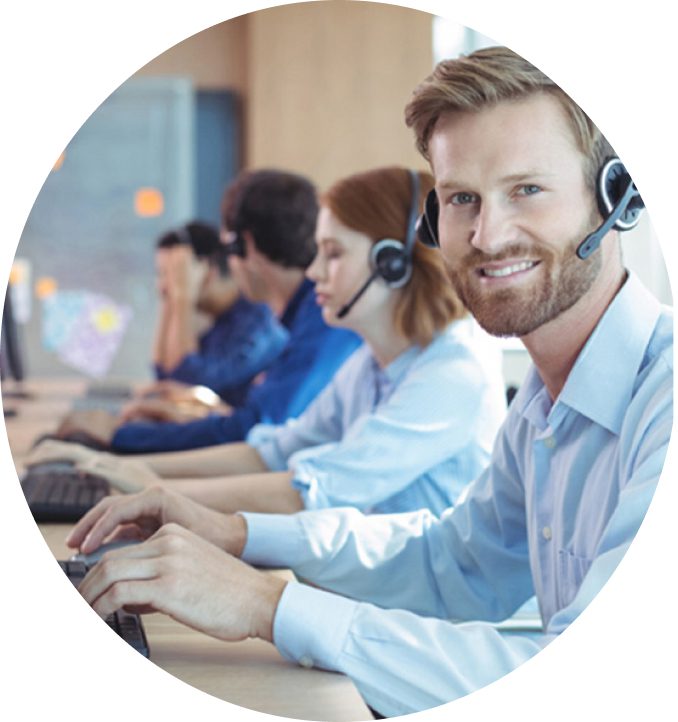 A imagem, em formato circular, mostra um grupo de pessoas em um ambiente de escritório, possivelmente um call center. Em primeiro plano, um homem sorri para a câmera enquanto digita em um teclado, usando um headset. Ao fundo, outras pessoas também estão trabalhando em computadores com headsets, indicando um ambiente de atendimento ou suporte.