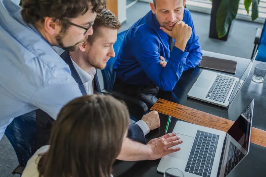 A imagem mostra um grupo de quatro pessoas em um ambiente de escritório, possivelmente em uma reunião ou sessão de trabalho colaborativo. Uma mulher de óculos em primeiro plano está focada em um laptop, enquanto um homem atrás dela parece estar olhando para uma tela ou tablet. Outro homem, de pé, também observa algo em um tablet, e uma mulher à direita está atenta ao que está acontecendo. O ambiente é bem iluminado por grandes janelas ao fundo, e a cena transmite uma atmosfera de produtividade e foco.