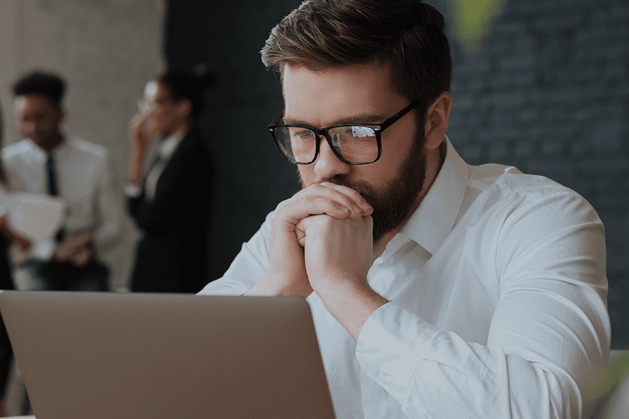 A imagem mostra um homem de barba e óculos, usando camisa social branca, concentrado em frente a um laptop. Ele está com as mãos unidas encostadas no rosto, em posição pensativa. Ao fundo, desfocados, aparecem duas pessoas em pé conversando, transmitindo o ambiente de escritório.
