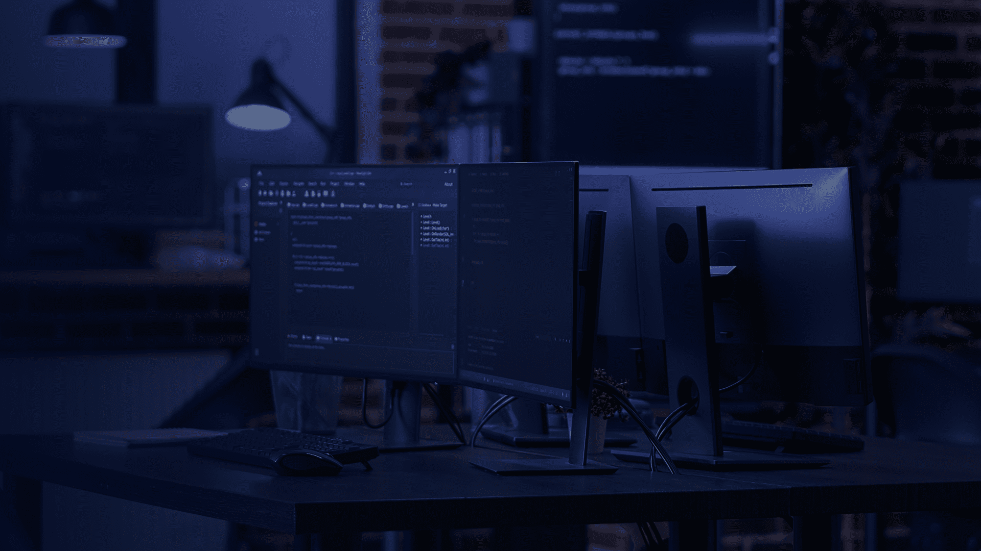 Sala escura com monitores ligados exibindo código, teclado e mouse sobre a mesa, em ambiente moderno de desenvolvimento de software.
