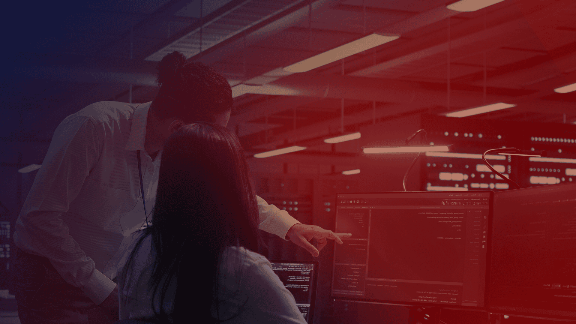 APIs.Homem em pé aponta para a tela e mulher sentada observa o código em um data center, com filtro azul e vermelho